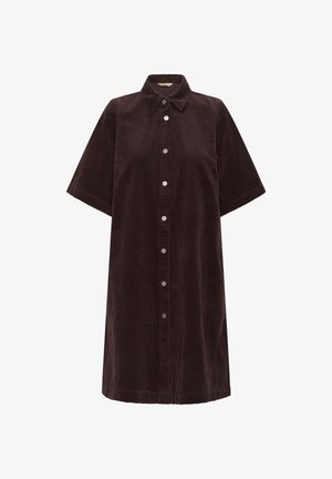 Kobieta ubrana w ciemnobrązową, sztruksową sukienkę na guziki o długości do kolan z krótkimi rękawami oraz czarne, sięgające kolan, botki na obcasie na jednolitym tle.