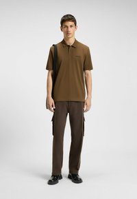 Brązowa koszulka polo z subtelnym logo, w połączeniu z ciemnobrązowymi spodniami cargo w wzory oraz czarnym obuwiem. Model stoi na jasnym tle.