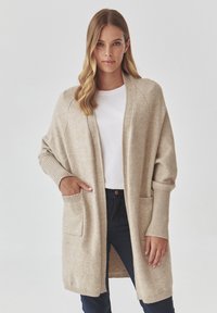 Cardigan beige à manches longues, avec poches avant et poignets côtelés. Texture douce, design ouvert, porté sur une chemise blanche.