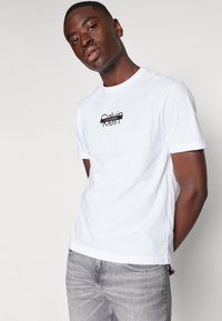 Hvit bomulls T-skjorte med kort erm, med en svart trykt Calvin Klein-logo på brystet. Kombinert med lysegrå jeans.