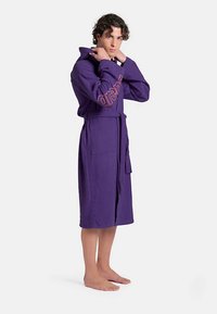 Peignoir violet en tissu doux, avec ceinture, poches latérales et un logo orange sur la poitrine. Sa longueur atteint le milieu du mollet.