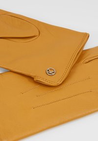 Gants en cuir jaune moutarde, avec un design épuré, un poignet replié, des détails de couture et un accent circulaire en métal logo.