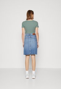 Camiseta verde de canalé combinada con una falda de mezclilla azul media. La falda cuenta con bolsillos traseros y una etiqueta de cuero marrón en la cinturilla.