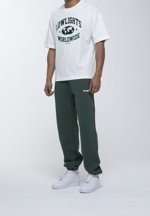 White short-sleeve t-shirt with green printed text and globe design; paired with dark green sweatpants and white sneakers.