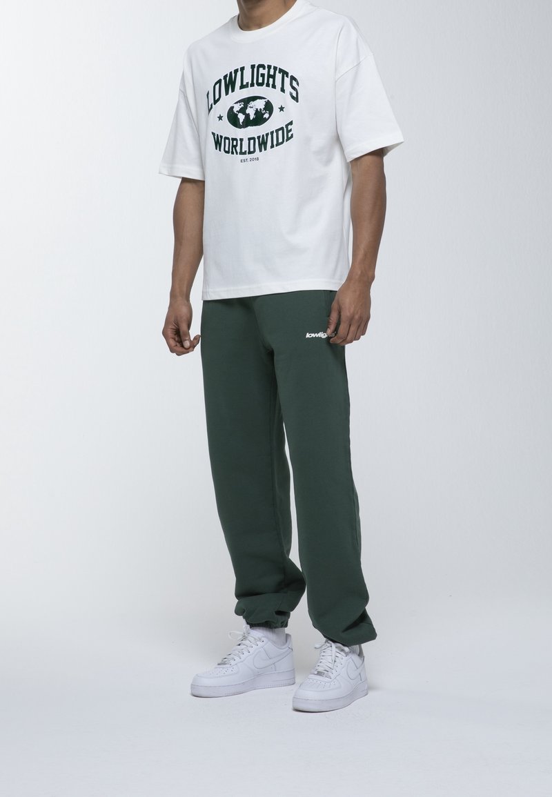T-shirt blanc à manches courtes avec texte imprimé vert et design de globe ; assorti à un pantalon de survêtement vert foncé et des baskets blanches.