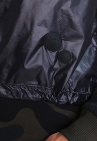 Acercamiento a la tela oscura y brillante de una chaqueta con dos botones a presión negros, uno grabado con "G RAW," sobre una prenda de patrón de camuflaje.