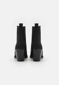 Botines de ante negro hasta el tobillo con tacón cuadrado, que cuentan con tiradores y detalles de costura vertical en los lados. Textura suave, diseño minimalista.