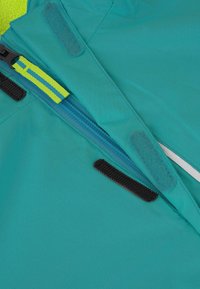 Türkiser Jackenstoff mit glatter Textur; verfügt über einen blauen Reißverschluss mit einer gelben Zuglasche und schwarzen Klettverschluss-Akzenten in der Nähe der Öffnung.