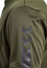 Olivgrön tyg med tre svarta chevronformer vertikalt placerade på en ärm, med synlig sömnad och textur.