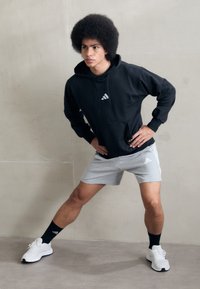 Joven con afro estirando las piernas, manos en las caderas, vistiendo sudadera negra con capucha, pantalones cortos grises, calcetines negros y zapatillas blancas frente a una pared lisa.