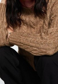 Pull en maille torsadée de couleur brun clair et chaud avec de larges poignets côtelés. Le tissu semble doux et texturé, associé à un pantalon noir.