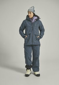 Navy waterproof jacket with two front pockets, layered over a light purple top, paired with navy pants and cream snow boots.