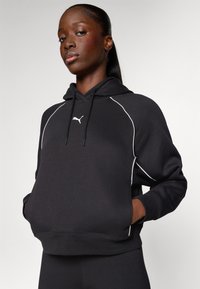 Svart hettegenser laget av myk stoff, med en hvit Puma-logo, kontrasterende hvite sømmer langs raglanermene, og en frontlomme.