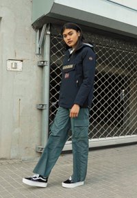 Marineblauwe jas met een ritssluiting aan de voorkant, een logo op de borst en vlaggen; groene cargobroek; zwart-witte sneakers; stedelijke achtergrond met een rasterhek.