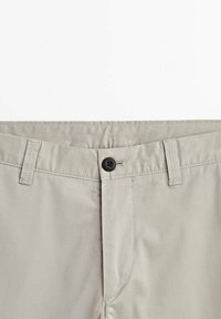 Taille de pantalon beige avec bouton noir, braguette zippée, passants de ceinture et coutures visibles sur fond blanc uni.