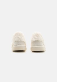 Hvide pelsforede slippers med en glat, blød tekstur, en overdel i ruskind og en lysegrå gummisål. Rundet bagdesign.