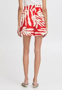 Žena v šortkách s vysokým pasem, červenobíle vzorovaných s abstraktním designem, spojených s bílými sandály na podpatku.