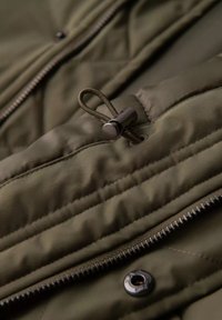 Olivově zelená prošívaná bunda s hladkým povrchem, která má šňůrku se stahovacími kovovými kuličkami a zipové zavírání s kovovým knoflíkem.