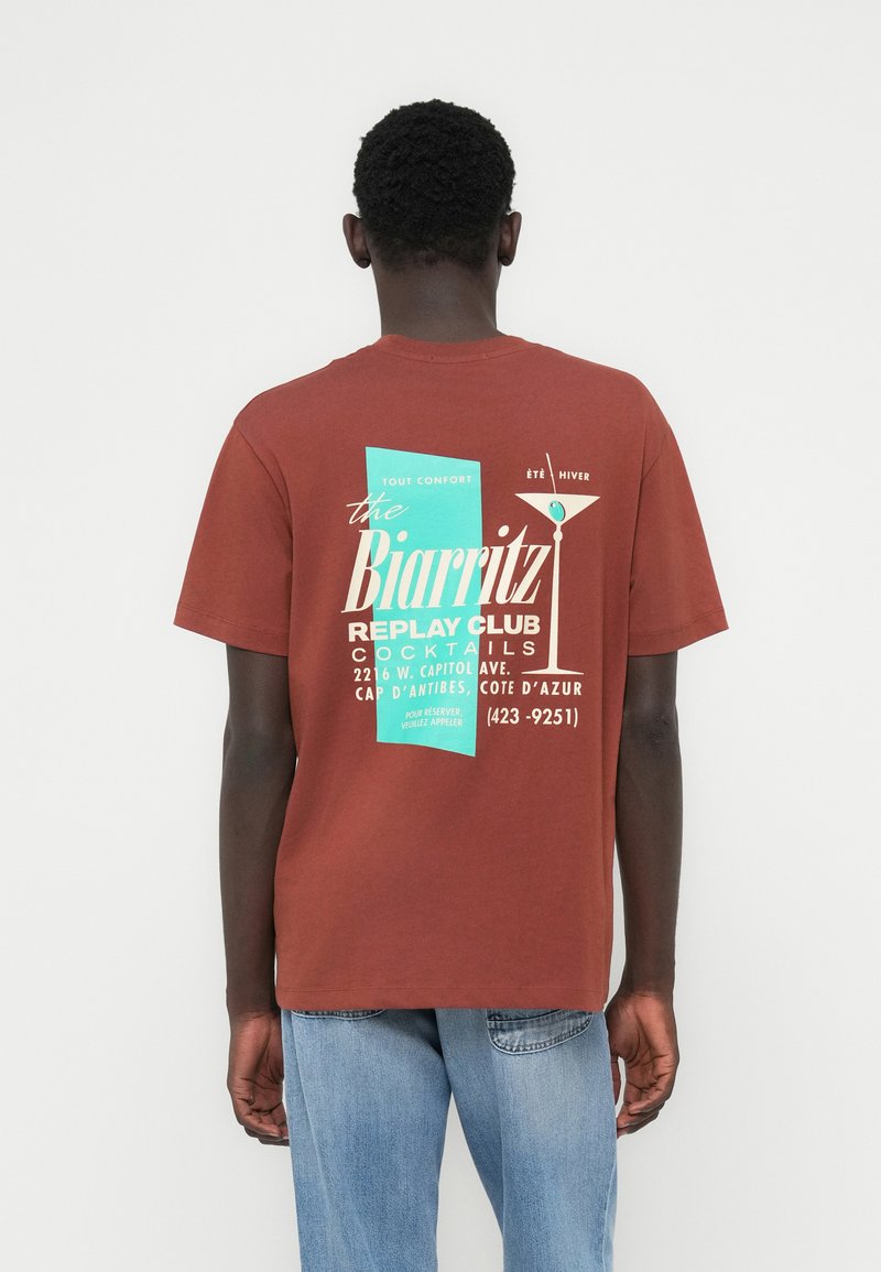 Καφέ βαμβακερό t-shirt με γραφικό σχέδιο στην πλάτη που περιλαμβάνει κείμενο σε τιρκουάζ και κρεμ αποχρώσεις, σχέδιο ποτηριού μαρτίνι και άνετη εφαρμογή.