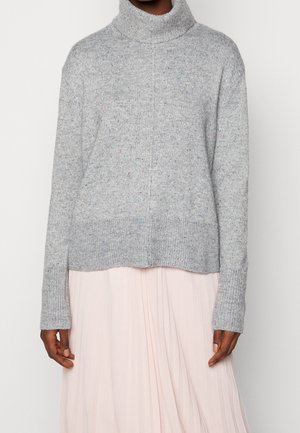 Personne portant un pull à col roulé gris clair à manches longues et une jupe plissée rose clair devant un fond uni.