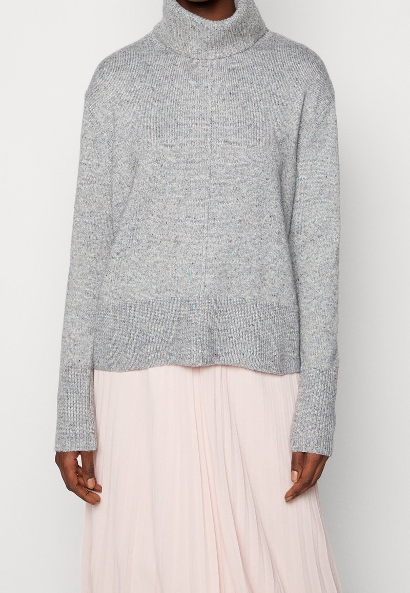 Personne portant un pull à col roulé gris clair à manches longues et une jupe plissée rose clair devant un fond uni.