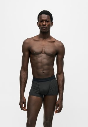 Atletisk mørk hudfarvet mand, der står vendt fremad iført tætsiddende sorte bokserbriefs mod en ensfarvet hvid baggrund.