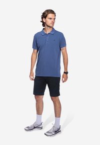 Mannen marineblauwe polo met een dubbele knoopsluiting, korte mouwen, gedragen met zwarte shorts, grijs-witte sportschoenen en lichte sokken.