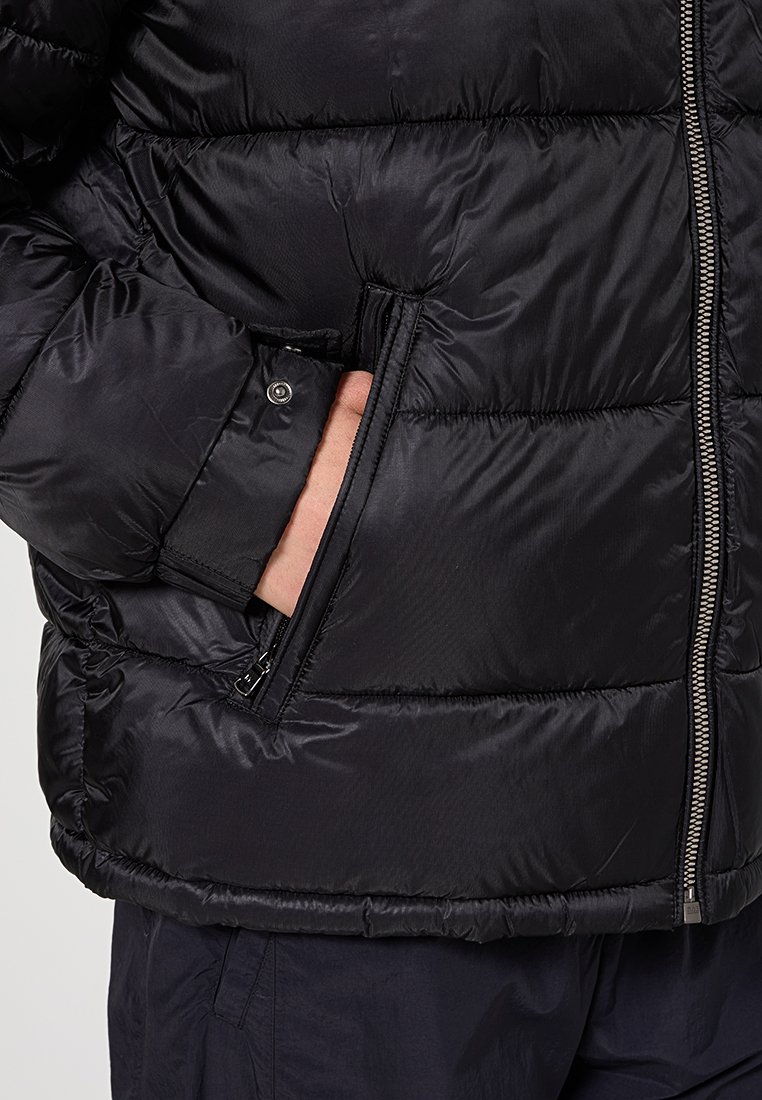 Giacca nera trapuntata con cuciture orizzontali, chiusura con zip e una mano inserita nella tasca laterale con zip.