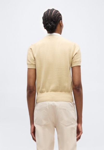 Άτομο με πλεξούδες που φοράει μπεζ πουλόβερ με κοντά μανίκια και ανοιχτόχρωμο παντελόνι, εμφανιζόμενο από πίσω σε απλό φόντο.