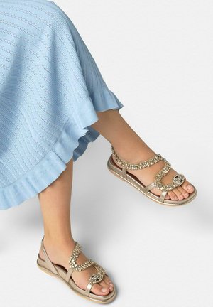 Piedi che indossano sandali gioiello dorati abbinati a una gonna azzurro chiaro con trama e orlo arricciato su sfondo bianco.