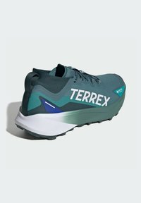Scarpe sportive in un verde scuro con il branding bianco "TERREX", tomaia in tessuto morbido, una suola spessa bianca e verde, e un battistrada dettagliato per la trazione.