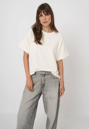 Kobieta o długich brązowych włosach, ubrana w luźną białą koszulę z krótkim rękawem i szerokie, jasnoszare dżinsy, stojąca na jednolitym białym tle.