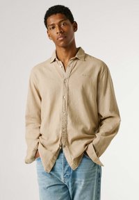 Νεαρός άνδρας που φοράει μπλούζα μπεζ με κουμπιά και μανίκια γυρισμένα, καθώς και ανοιχτόχρωμο μπλε τζιν, στέκεται με τα χέρια στις τσέπες μπροστά σε απλό φόντο.