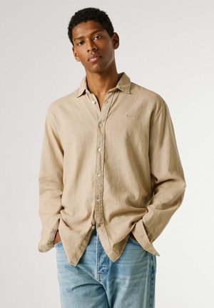 Mladi moški, oblečen v bež srajco z dolgimi rokavi, zavihani manšeti, in svetlo modre kavbojke, stoji z rokami v žepih pred enobarvnim ozadjem.