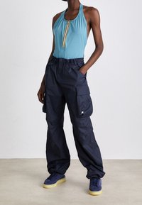 Personne portant un haut bleu sans manches à fermeture éclair, un pantalon cargo noir ample avec des poches latérales, et des baskets bleues avec des semelles jaunes.