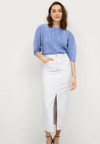 Blaues, gefaltetes Oberteil mit weiten Ärmeln, kombiniert mit einem weißen, hoch taillierten Jeansrock mit einem Schlitz vorne. Schlichtes, sauberes Design.