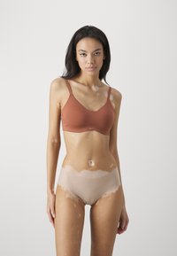 Smeđa bralette s podesivim trakama i bešavnim dizajnom, uparen s bež bešavnim gaćicama. Ova dva komada imaju glatku teksturu.