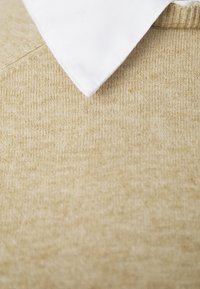 Beżowy sweter dzianinowy o teksturowanym wykończeniu z widoczną białą bawełnianą kołnierzykiem przy dekolcie.