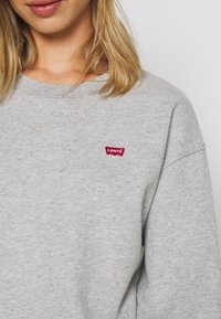 Sweat-shirt en polaire grise avec un col rond, des manches longues et un patch logo rouge Levi's sur la poitrine gauche. Texture douce avec un léger effet chiné.
