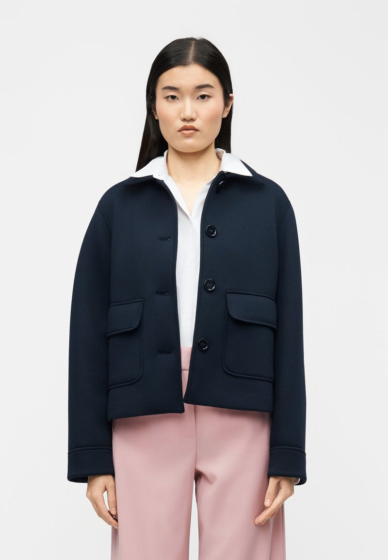 Γυναίκα με ίσια μαύρα μαλλιά φοράει ένα σκούρο μπλε σακάκι με κουμπιά πάνω από λευκό πουκάμισο και ροζ παντελόνι με φαρδιά πόδια, στέκεται μπροστά σε απλό φόντο.