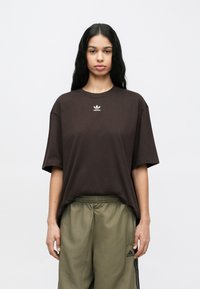 Νέος με μακριά μαύρα μαλλιά, φορώντας μια χαλαρή σκούρη καφέ μπλούζα Adidas και χακί παντελόνι φόρμας Adidas, στέκεται μπροστά σε ένα απλό φόντο.