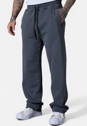 Mann trägt dunkelgraue Jogginghose mit Kordelzug und weiße Sneakers, die Hände ruhen in den Taschen, sichtbare tätowierte Unterarme.