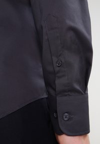 La maglietta grigia scura a maniche lunghe presenta un tessuto liscio, polsini con bottoni composti da due bottoni e un design ben sartoriale.