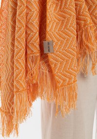 Oranje geweven shawl met een vissengraatpatroon, franjes aan de randen en een klein stoffen label. Vergezeld door een lichtgekleurde stoffen achtergrond.