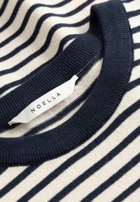 Posnetek v bližini modro-belih črtastih pletenih roba puloverja z belo etiketo, na kateri je izpisano ime znamke "NOELLA".