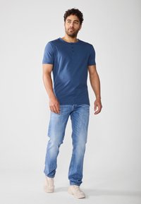 Maglietta henley a maniche corte blu navy con placca a tre bottoni, abbinata a jeans in denim azzurro chiaro e sneakers color beige chiaro.