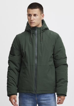 Zeleni puffer jakna sa patentnim zatvaračem sprijeda, kapuljača sa utezima i kviltanom teksturom. Ima raglan rukave i ravni rub. Uparena s plavim trapericama.