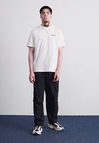 Hvid bomulds T-shirt med lille logo, sorte afslappede bukser og sort-hvide sneakers. Ensfarvet baggrund fremhæver fokus på tøjet.