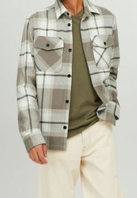 Camicia a quadri marrone e bianca con bottoni neri e due tasche sul petto, vestibilità comoda, indossata sopra una t-shirt verde oliva.