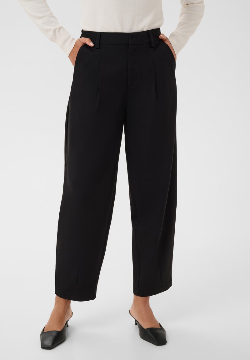 Femme portant un pantalon noir taille haute à jambes larges associé à des chaussures noires à bout pointu et talon bas, mains dans les poches, haut blanc à manches longues partiellement visible.